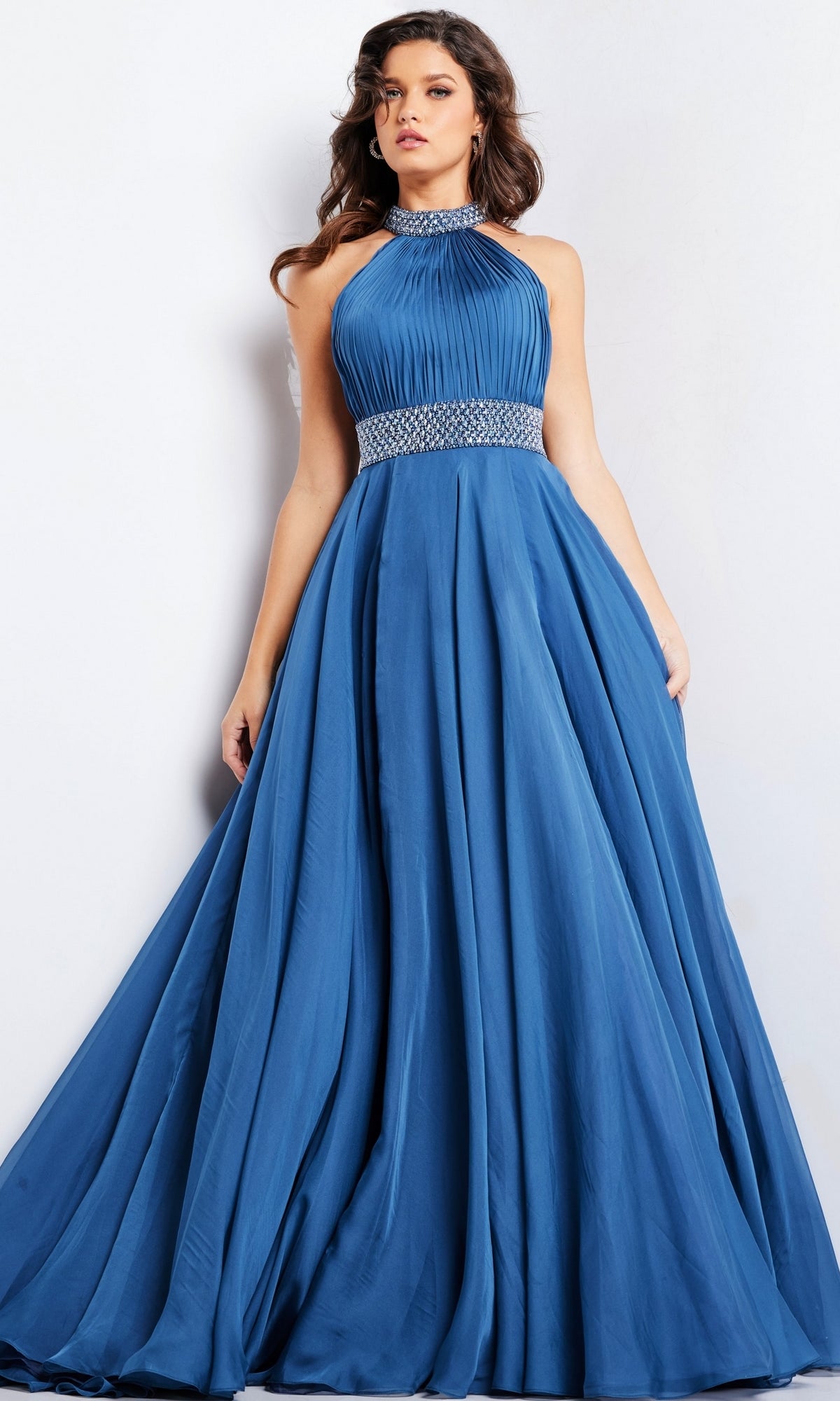 A woman stands against a plain light background, wearing the Long Prom Dress 36749 by Jovani—a sleeveless, floor-length a-line gown with a pleated bodice, beaded waistband, and flowing skirt.